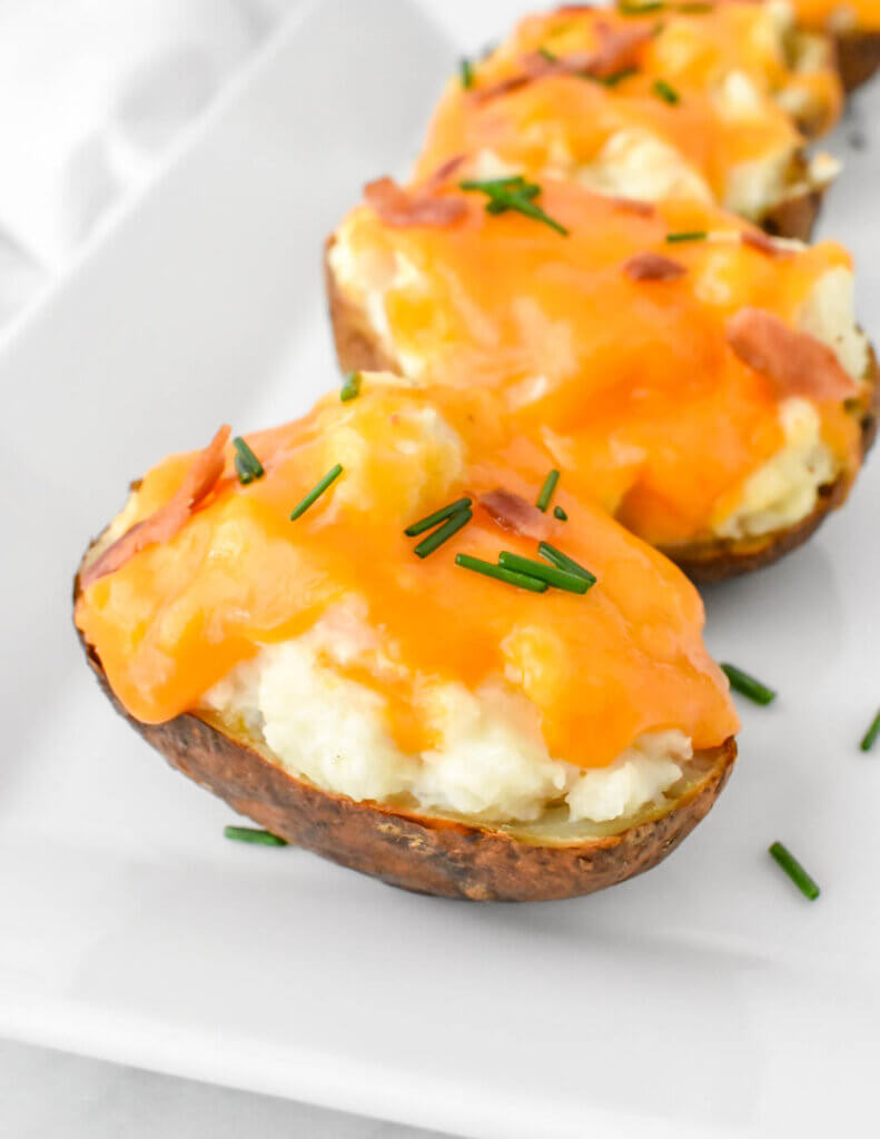 Twice baked potatoes topped with melted cheese, bacon and chives on a white rectangular platter.