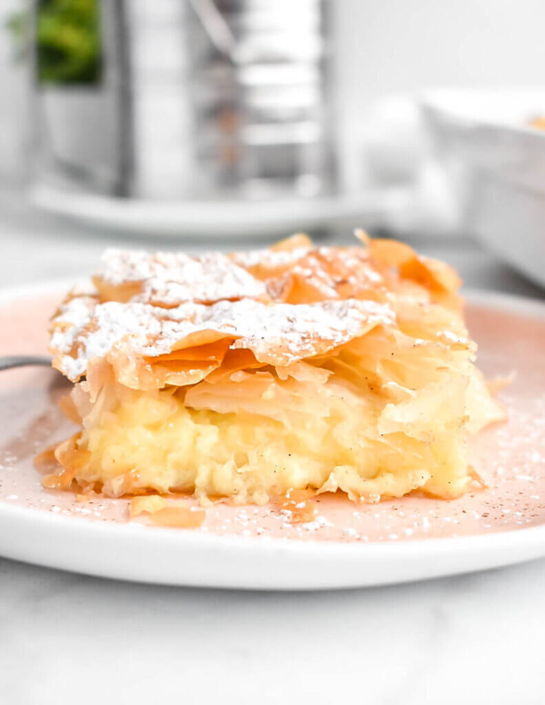 Bougatsa slice showing crispy filo and creamy custard layers on a pink plate.