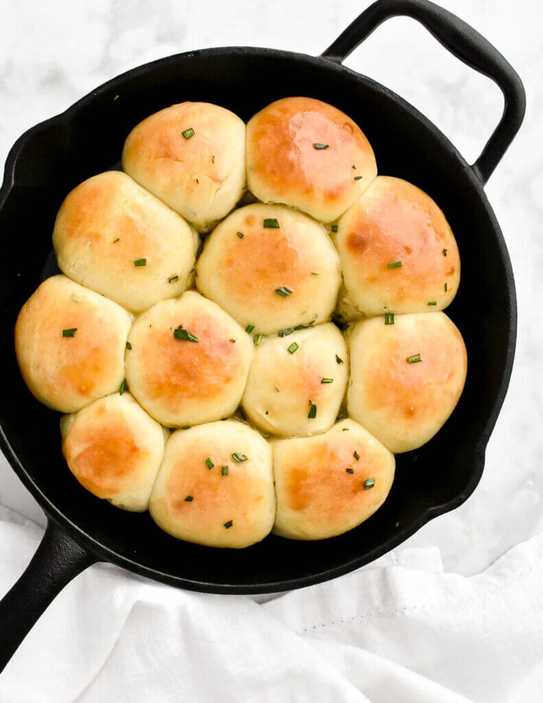 A cast iron skillet with baked dinner rolls topped with rosemary set on a white kitchen towel.