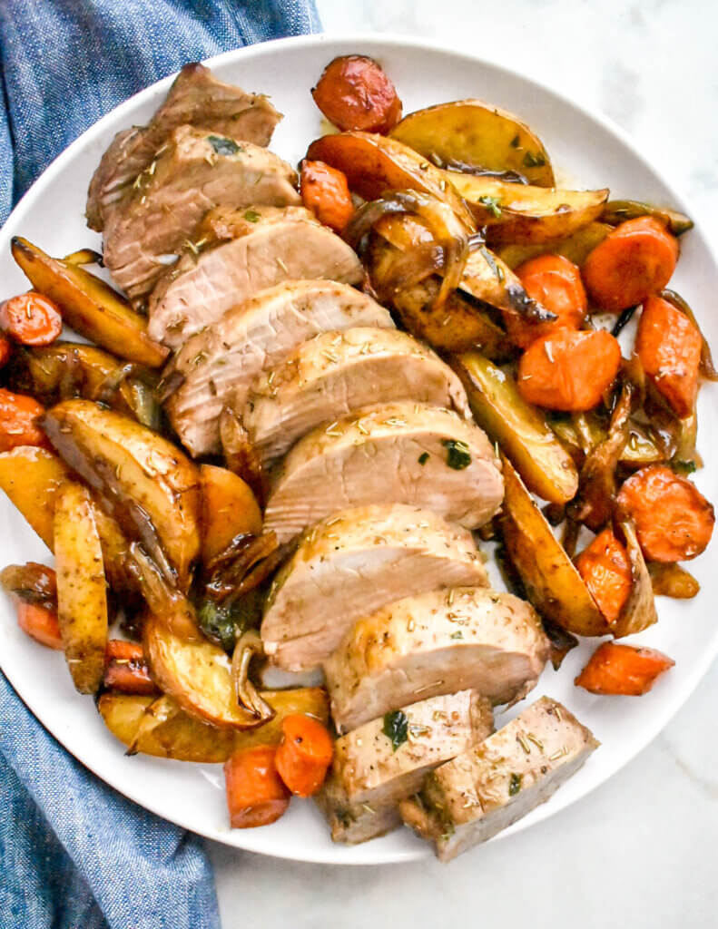 A round white serving platter with sliced pork tenderloin surrounded by roasted potatoes and carrots.