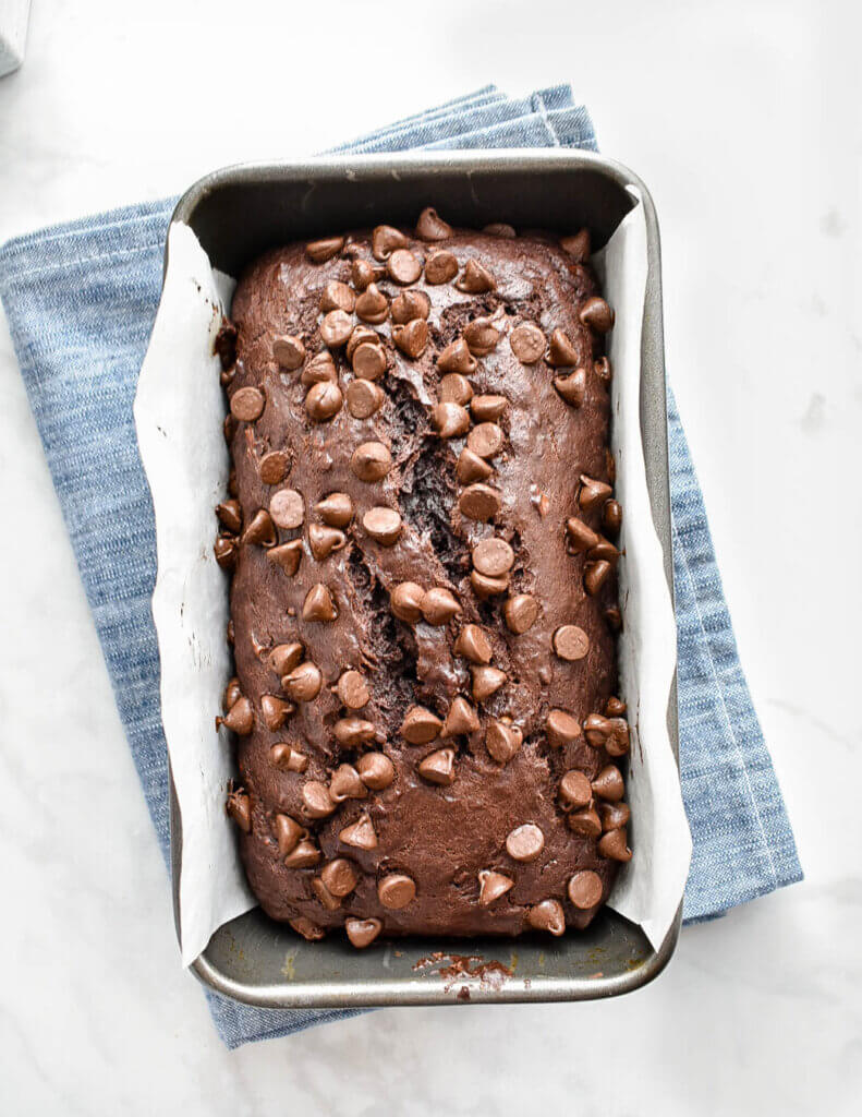 Baked Chocolate Banana Bread topped with chocolate chips in a loaf pan set on a blue kitchen towel.