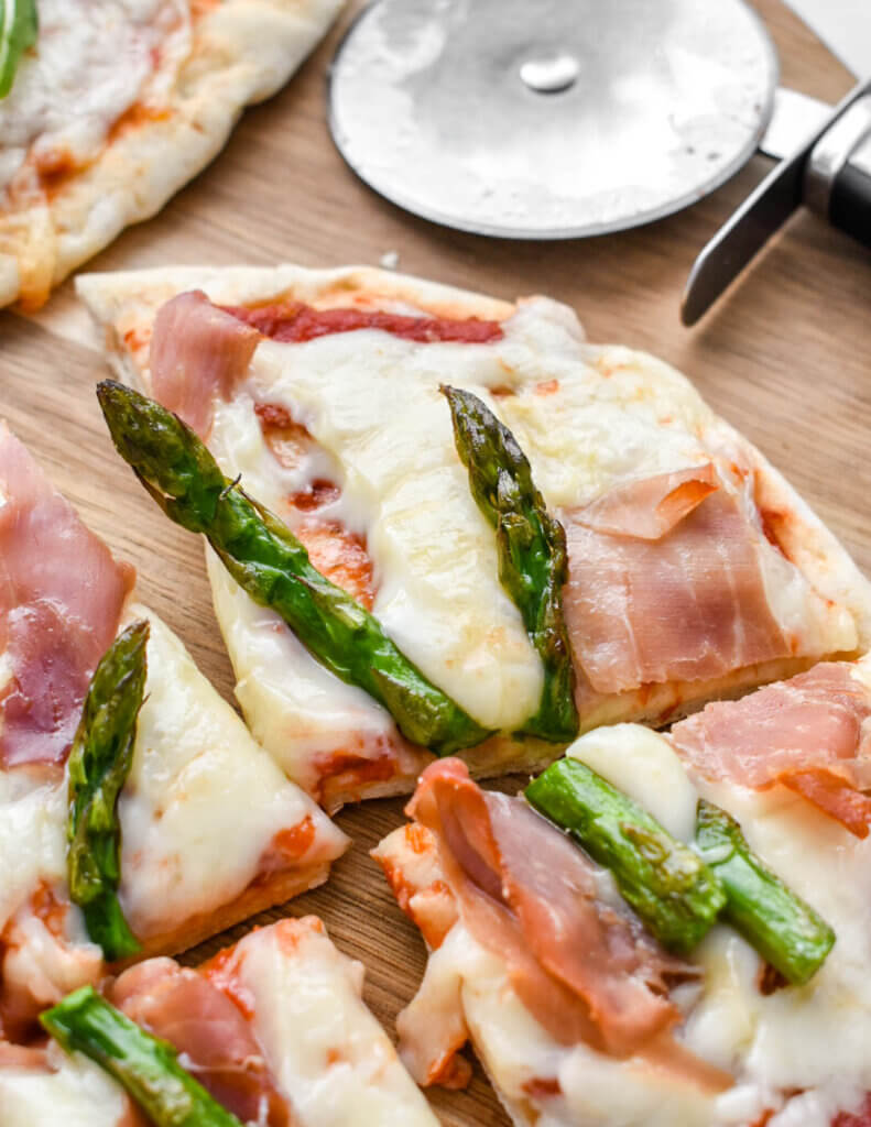 Closeup of a sliced pita pizza topped with asparagus and prosciutto on a cutting board next to a pizza cutter.