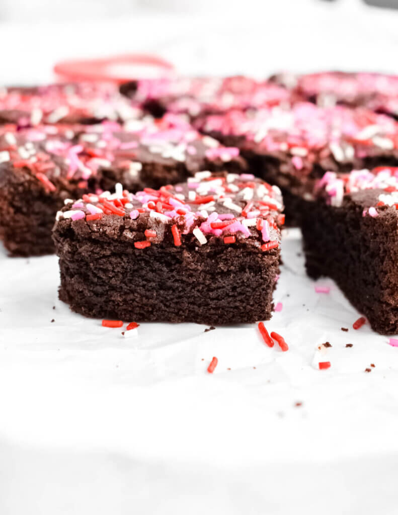 A heart shaped brownie covered in pink, red and white sprinkles on a parchment paper with other brownies.