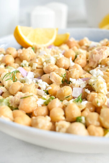 Closeup of chickpea salad with flakes of tuna, red onion, feta and fresh dill served with lemon wedges.