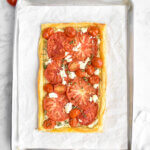 A tomato puff pastry tart made with heirloom tomato slices and halved grape tomatoes on a baking sheet lined with parchment paper.