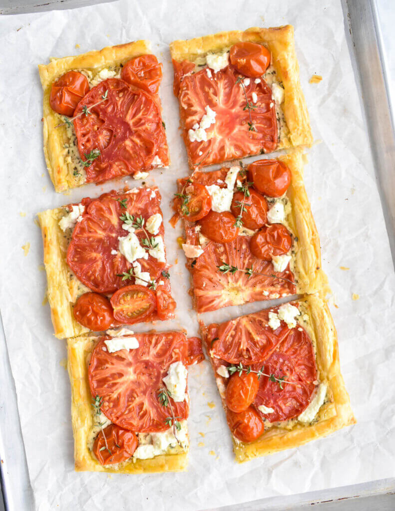 A sliced puff pastry tomato tart topped with goat cheese and fresh herbs on a baking sheet lined with parchment.