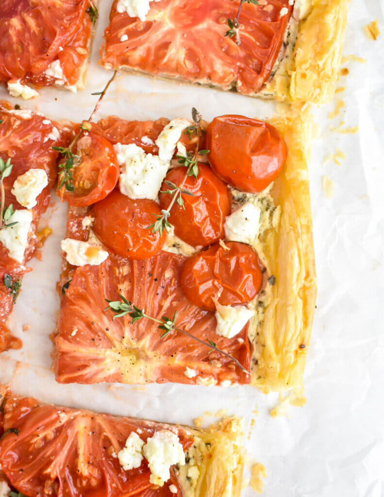 A closeup of a slice of puff pastry tart topped with halved grape tomatoes, heirloom tomato slices, crumbled goat cheese and fresh thyme.