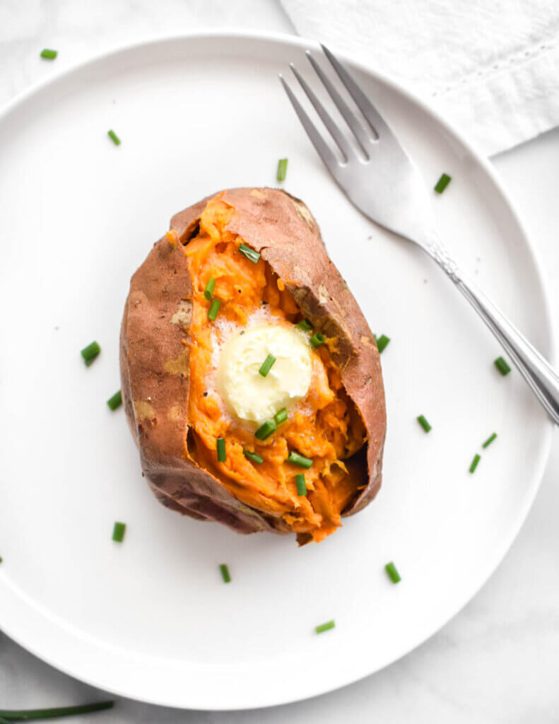 An air fryer baked sweet potato topped with butter and chives on a white plate.