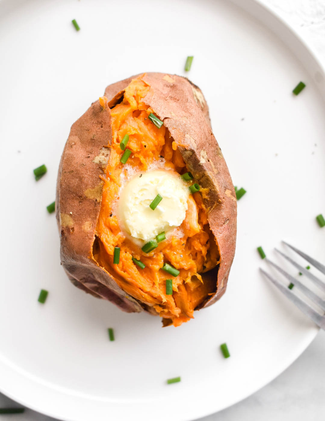 A baked sweet potato topped with a pat of butter and fresh chives on a white plate.
