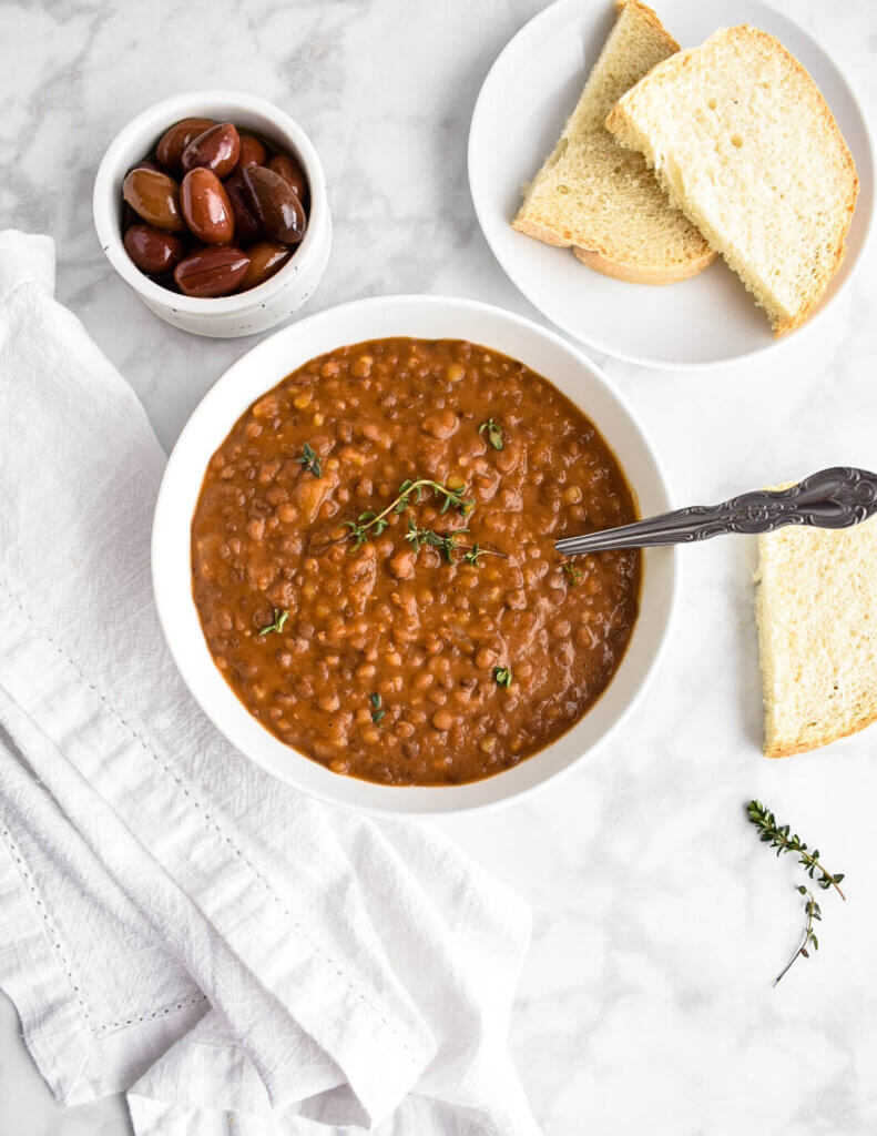 A bowl of fakies (Greek Lentil Soup) set on a marble countertop with a plate of sliced bread and a bowl of kalamata olives.
