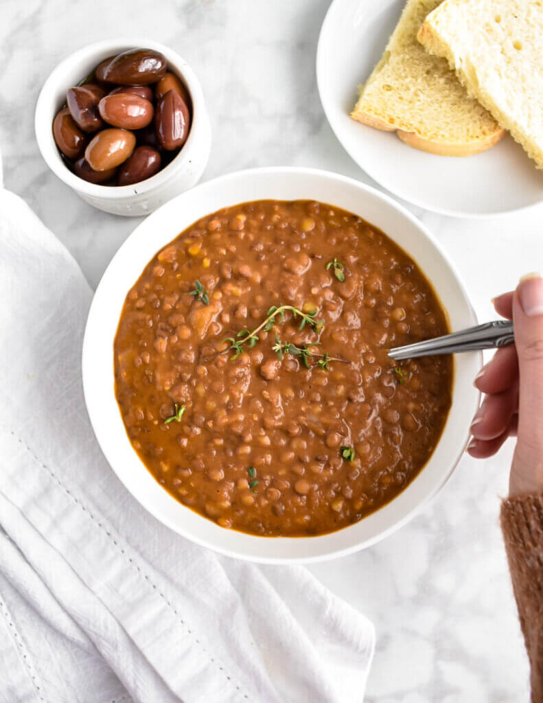 A bowl of lentil soup topped with thyme on a table next to olives and sliced bread.