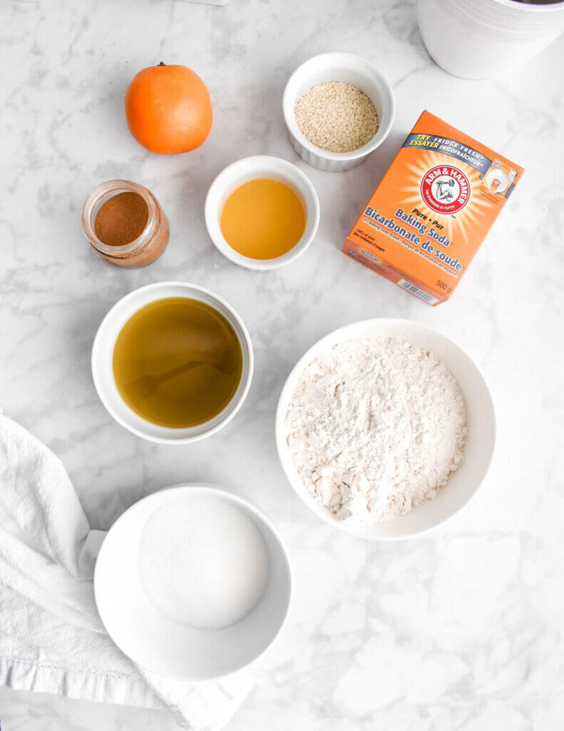 Bowls of flour, sugar, olive oil, orange juice, cinnamon, sesame seeds and a box of baking soda and whole orange on a marble countertop.