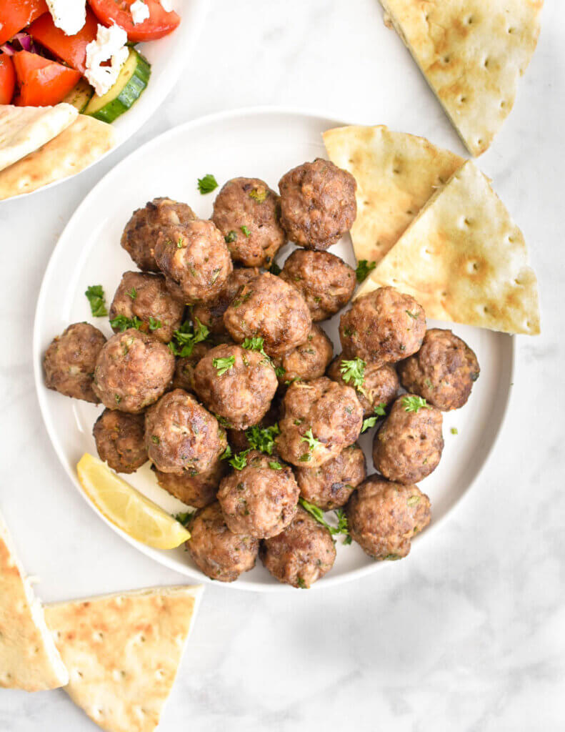 A platter of cooked meatballs topped with parsley on a kitchen counter surrounded by pita bread wedges.