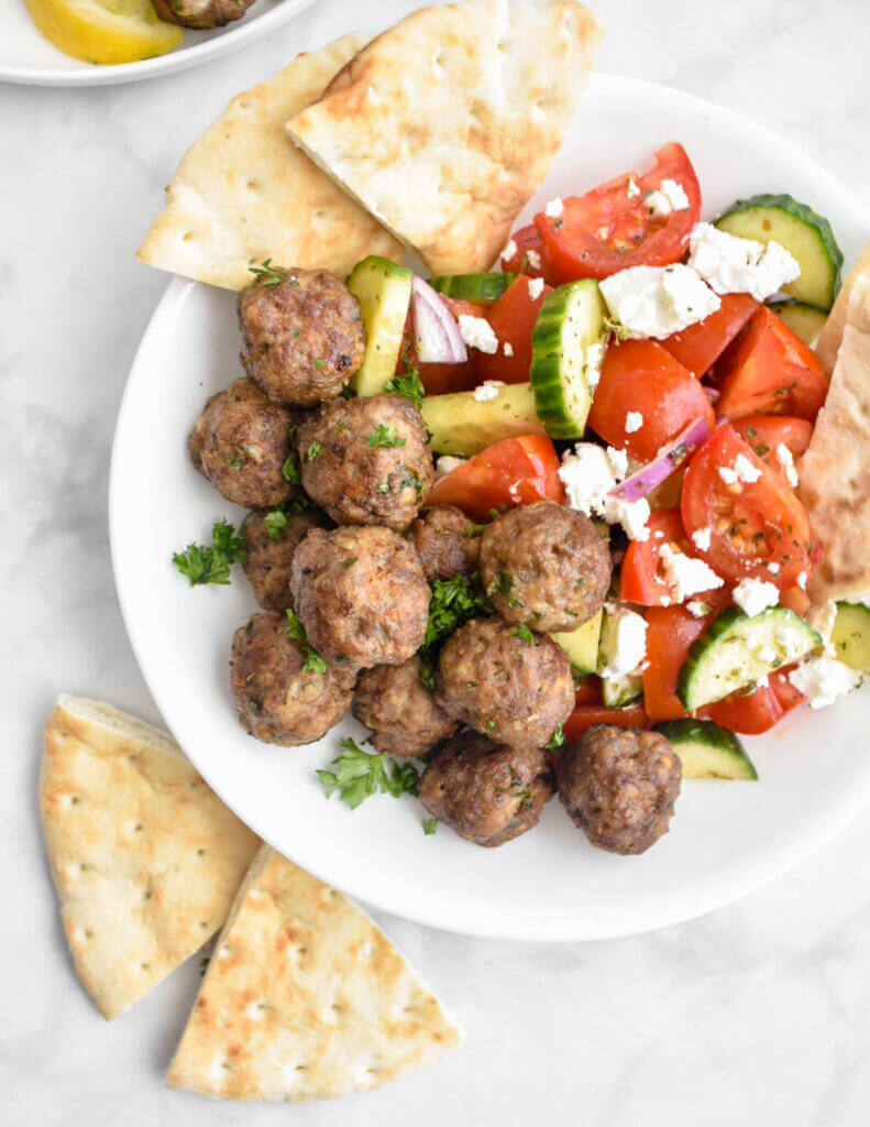 A white bowl filled with Greek Meatballs and Greek salad and pita wedges.
