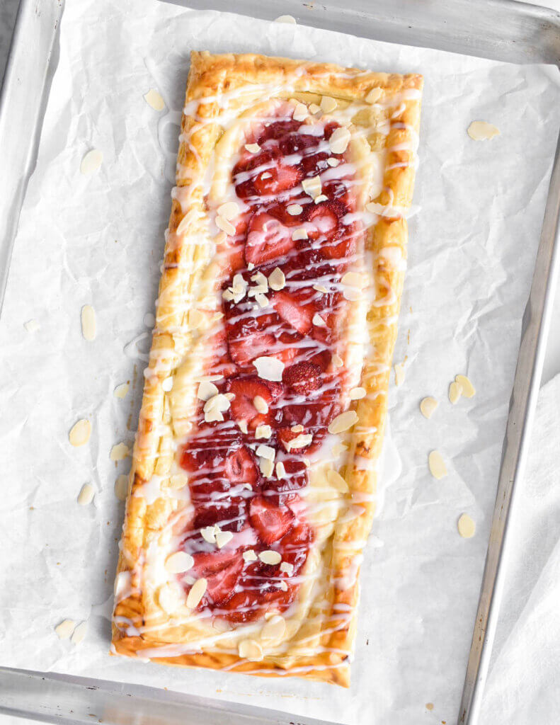 A baked strawberry cream cheese danish topped with icing and almond slivers on a parchment lined baking sheet.