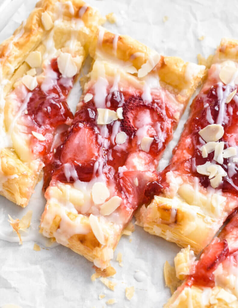 Close up of a sliced strawberry cream cheese danish showing the sliced strawberry and jam topping drizzled with icing and sliced almonds.