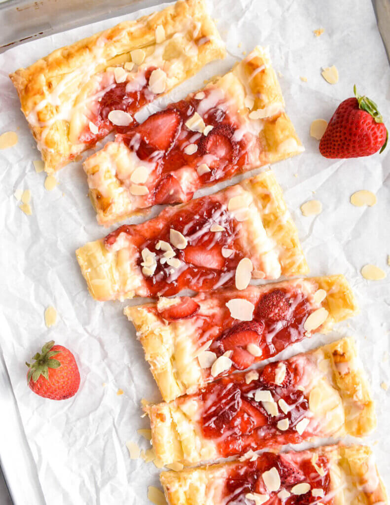 A long rectangular strawberry cream cheese danish that is drizzled with icing and sliced almonds and sliced into 6 portions.