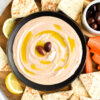 A bowl of taramosalata dip topped with olive oil and olives on a wooden platter surrounded by pita bread wedges.