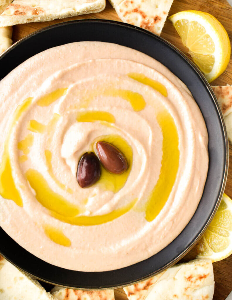 Closeup of a bowl of light pink taramosalata drizzled with olive oil and topped with black olives surrounded by pita and lemon wedges.