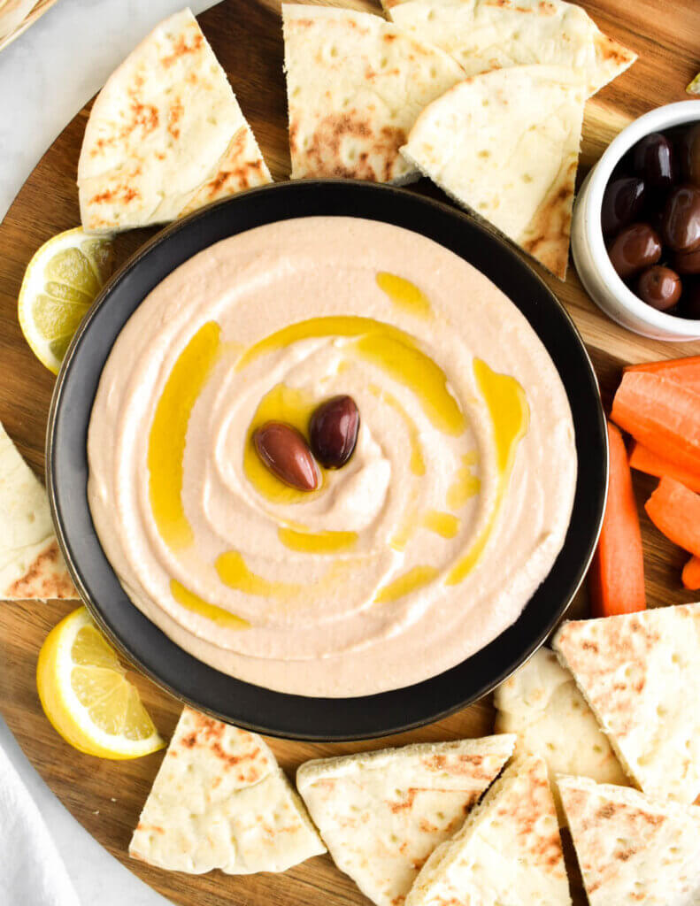 A bowl of taramosalata dip topped with olive oil and olives on a wooden platter surrounded by pita bread wedges.