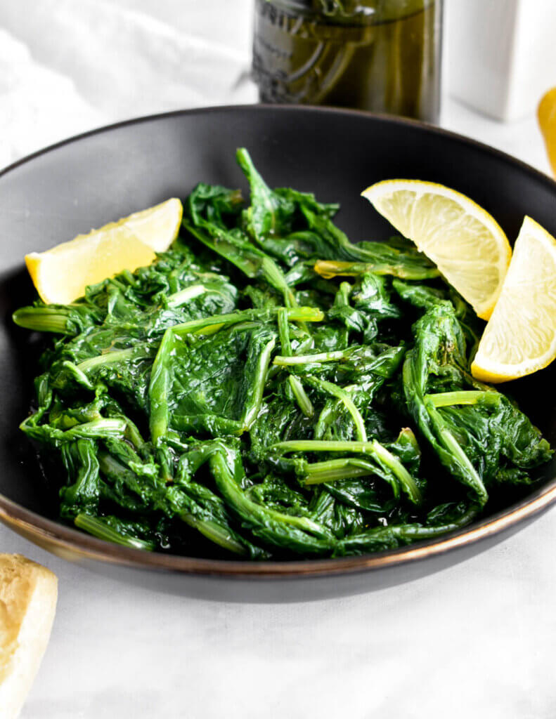 Boiled leafy greens in a black bowl served with lemon wedges.