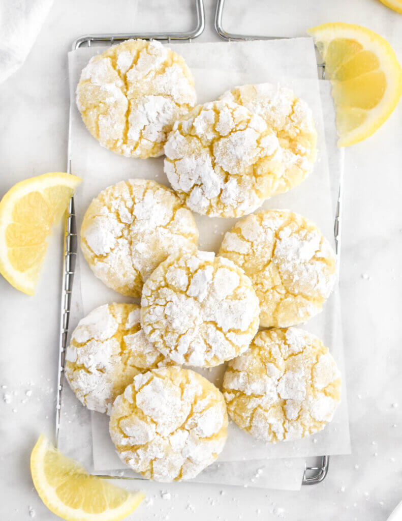Lemon Crinkle Cookies stacked on a small silver cooling rack surrounded by lemon wedges.