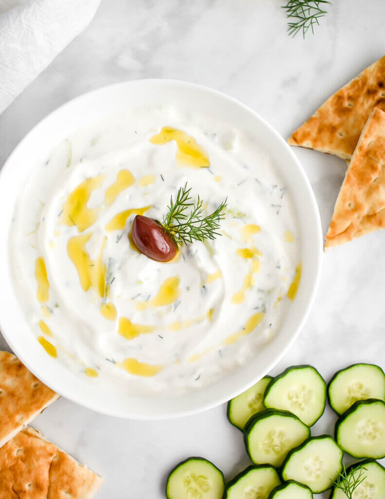 A bowl of tzatziki sauce topped with olive oil and an olive served with cucumber and pita slices.