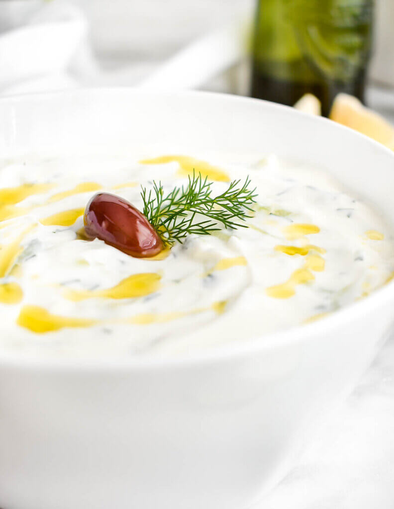 Closeup of a bowl of tzatziki drizzled with olive oil and topped with dill and an olive.