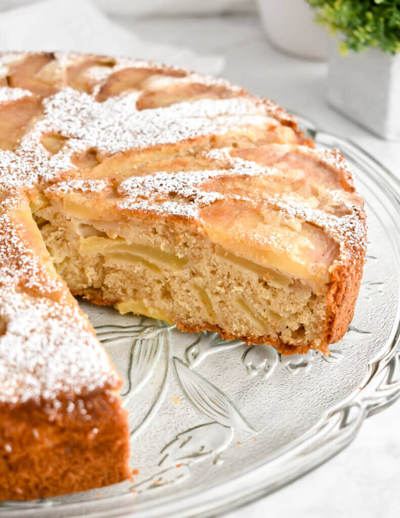 A sliced apple cake dusted with icing sugar on a clear platter.