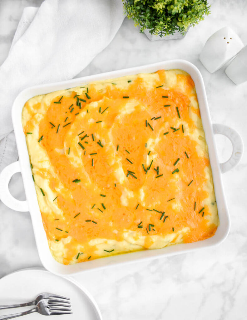 Baked mashed potatoes with a topping of melted cheddar cheese and chives in a square baking dish.