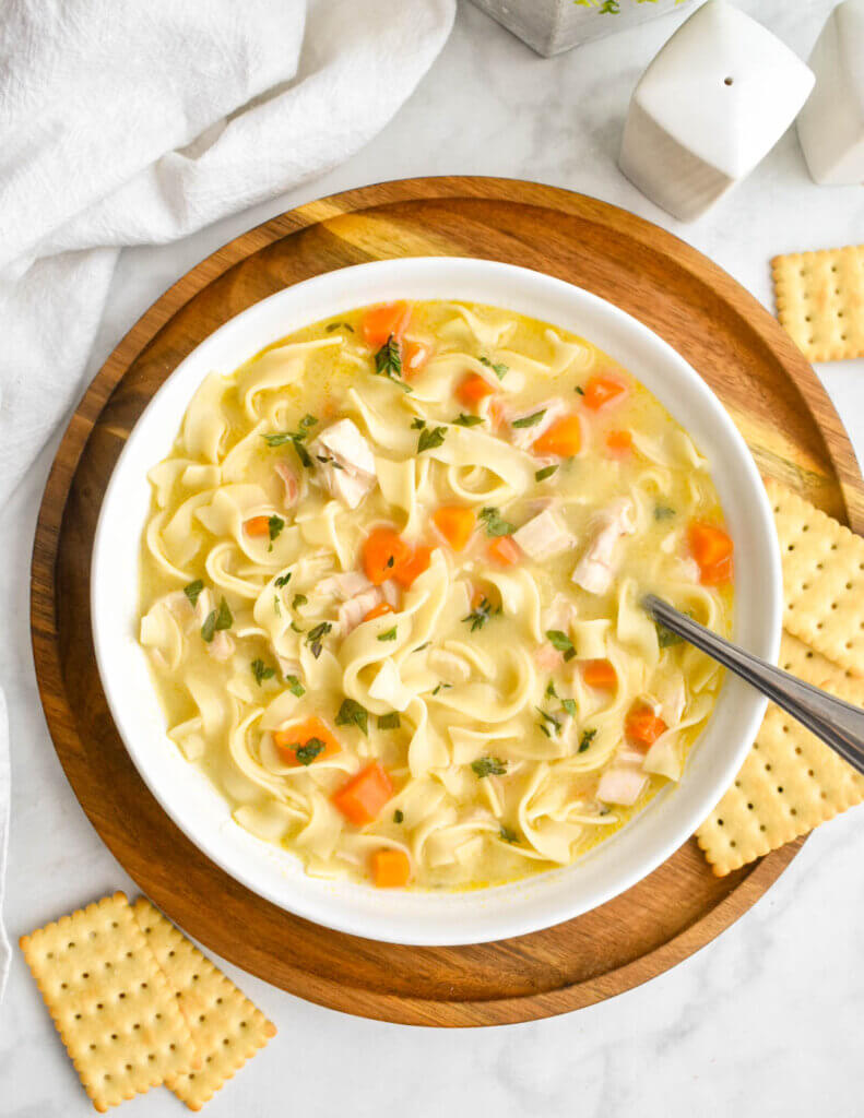 A bowl of chicken noodle soup set on a wooden charger plate and surounded with crackers, a white napkin and white salt and pepper shakers.
