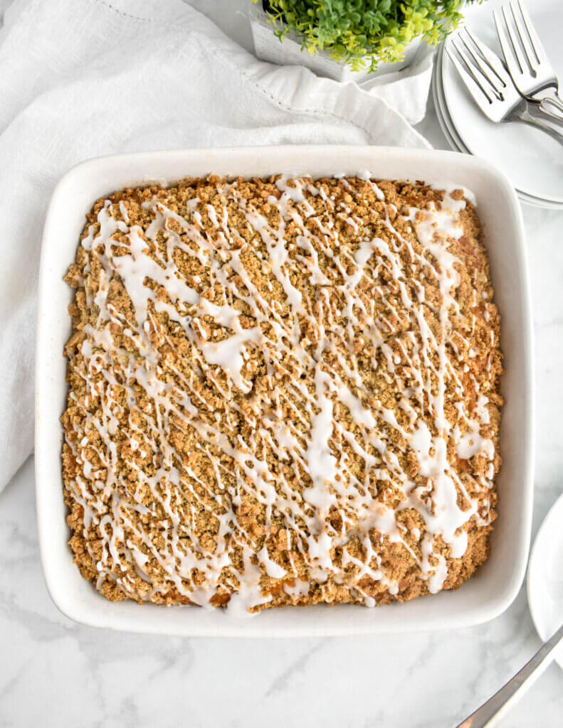 Top view of a coffee cake topped with crumb topping and a drizzled with icing in a zigzag pattern set on a grey marble countertop and surrounded by a white napkin and plates with forks.