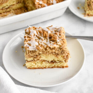 A slice of coffee cake with a crumb topping and icing drizzle on a white plate set down next to a white baking dish with coffee cake and a white cloth napkin in the foreground.