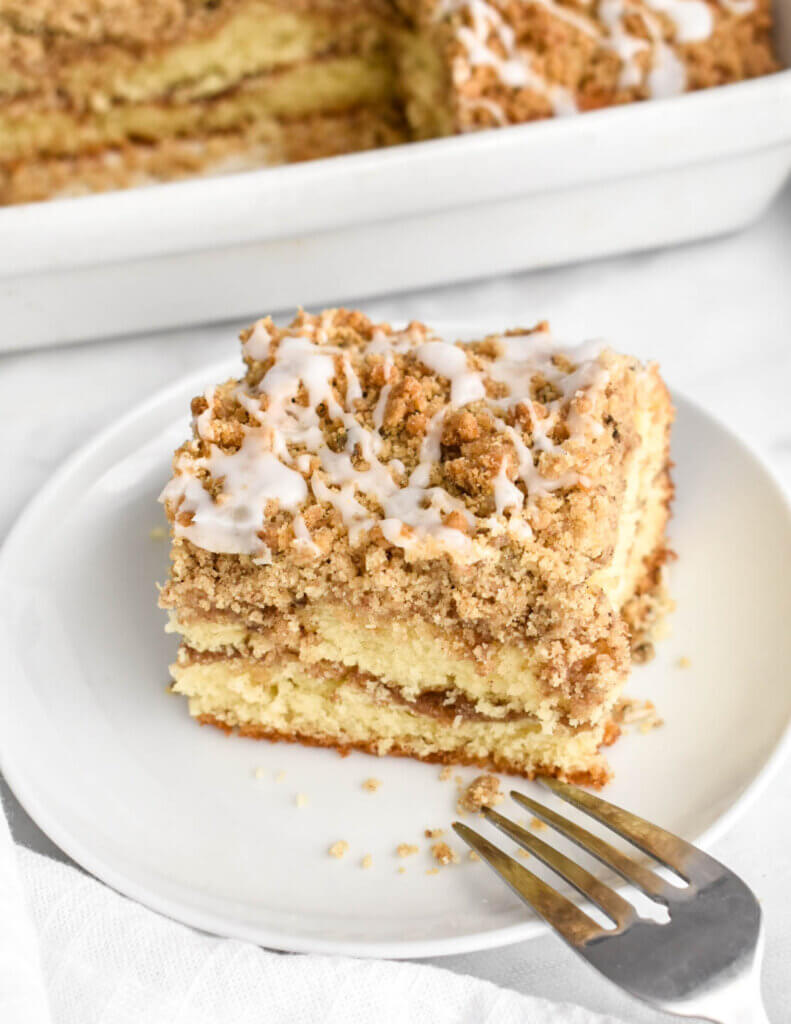 A slice of coffee cake with a cinnamon-sugar ribbon running through it a crumb topping with icing drizzle set on a white plate with a silver fork.