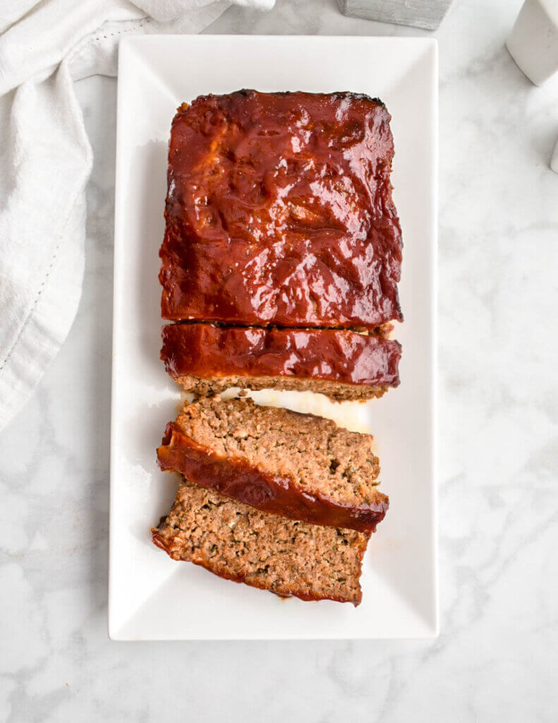 A meatloaf with a couple slices pre-cut on a white rectangular platter set on a grey marble countertop with a white napkin beside it.