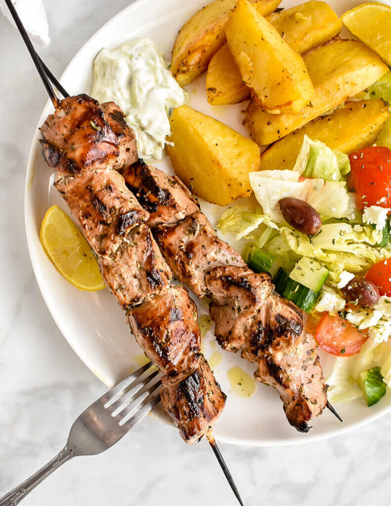 Closeup of two pork souvlaki skewers set on a white plate with a lemon wedge, tzatziki sauce, Greek salad and roasted potatoes.