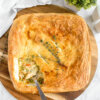 A Puff Pastry Turkey Pot Pie in a square baking dish set on a round wooden cutting board with a scoop of filling on a spoon.