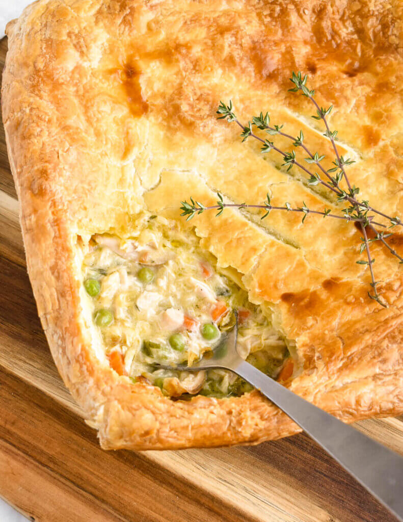 Closeup of a square turkey pot pie with the corner of pastry removed to show the creamy turkey filling being scooped up by a serving spoon.