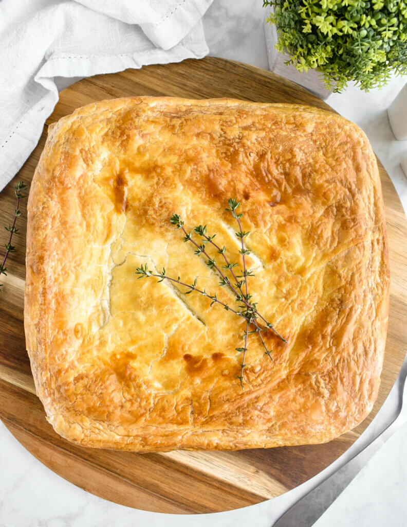 A square pan of turkey pot pie topped with puff pastry and garnished with thyme sprigs set on a wooden cutting board with a white napkin and green plant surrounding it.