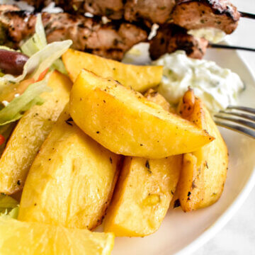 Crispy greek lemon potatoes on a plate with salad, pork souvlaki, tzatziki sauce and a lemon wedge.