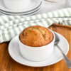 A single serve banana bread in a ramekin set on a white dish with a spoon next to it which in turn is set on a wooden cutting board with a second ramekin of banana bread showing in the background and a green and white striped napkin.