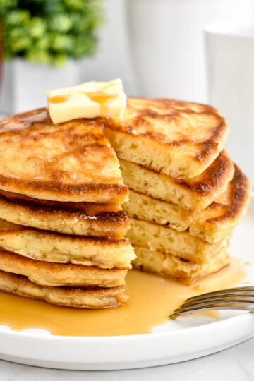 A stack of buttermilk pancakes topped with a pat of butter and maple syrup and cut into to reveal the fluffy interior.