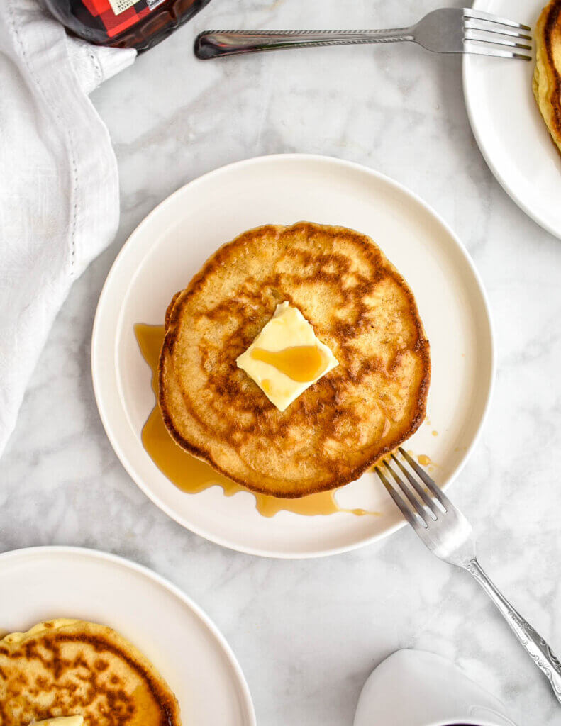 Top view of a plate of pancakes topped with syrup and butter and set on a white plate with a fork on a grey marble countertop with a white napkin, 2 other plates of pancakes, and a white mug of coffee set around it.