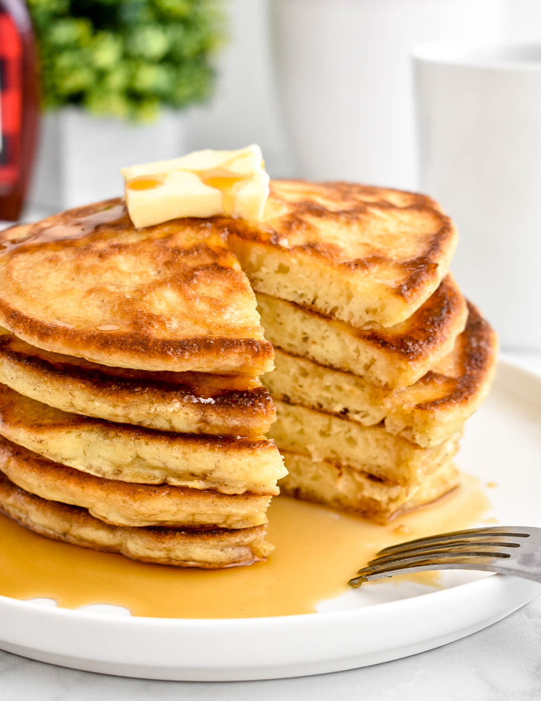 A stack of buttermilk pancakes topped with a pat of butter and maple syrup and cut into to reveal the fluffy interior.