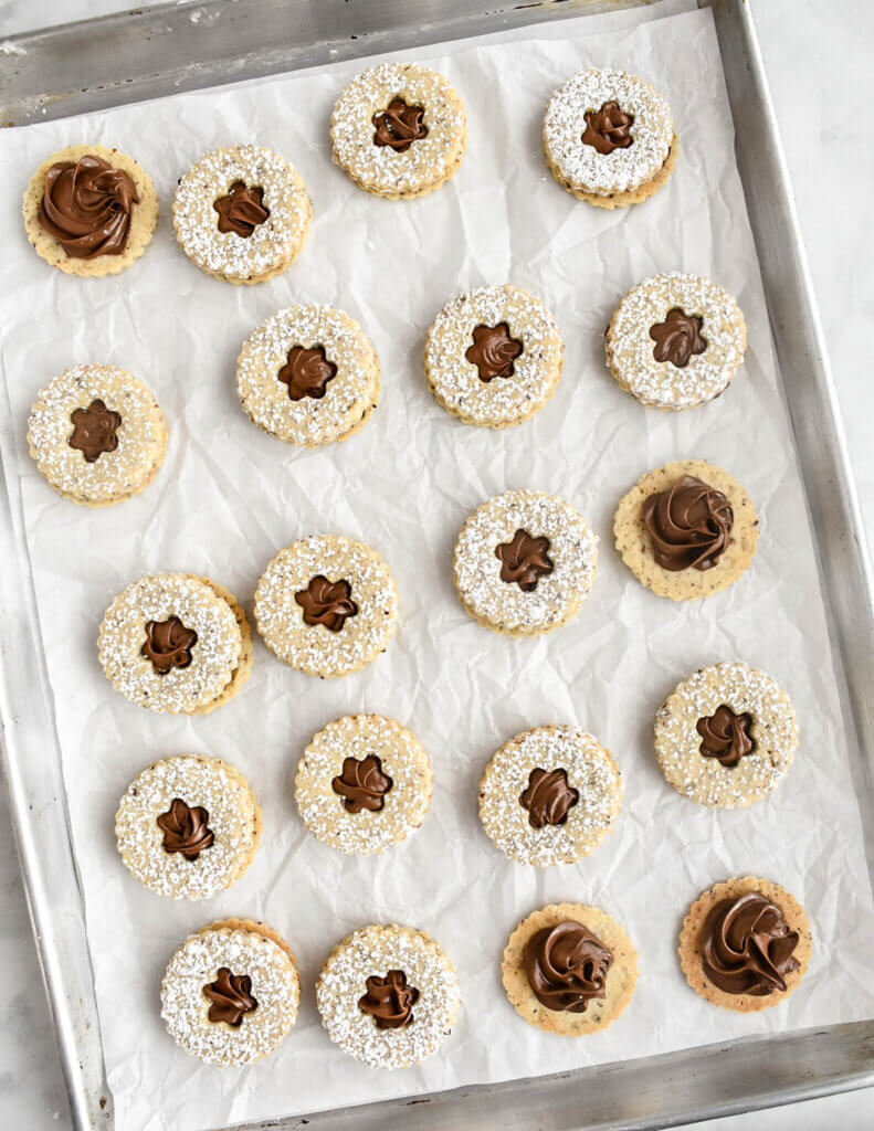 A tray of hazelnut cookie sandwiches with a flower shaped cut out showing the nutella filling on a baking tray with some cookies left untopped revealing the nutella inside.