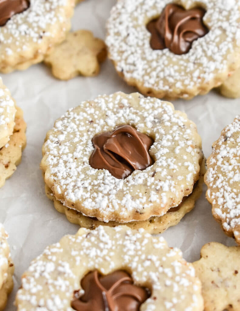 Closeup of a chocolate hazelnut linzer cookie with a flower shaped cutout on parchment paper surrounded by more cookies.