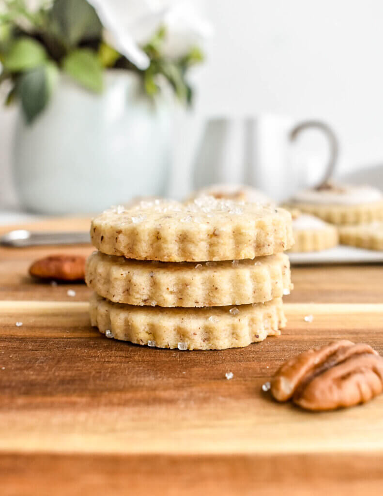 Three pecan cookies topped with coarse sugar stacked on top of each other set on a wooden cutting board with pecan halves next to them and a teal vase of white flowers and a coffee mug in the background.