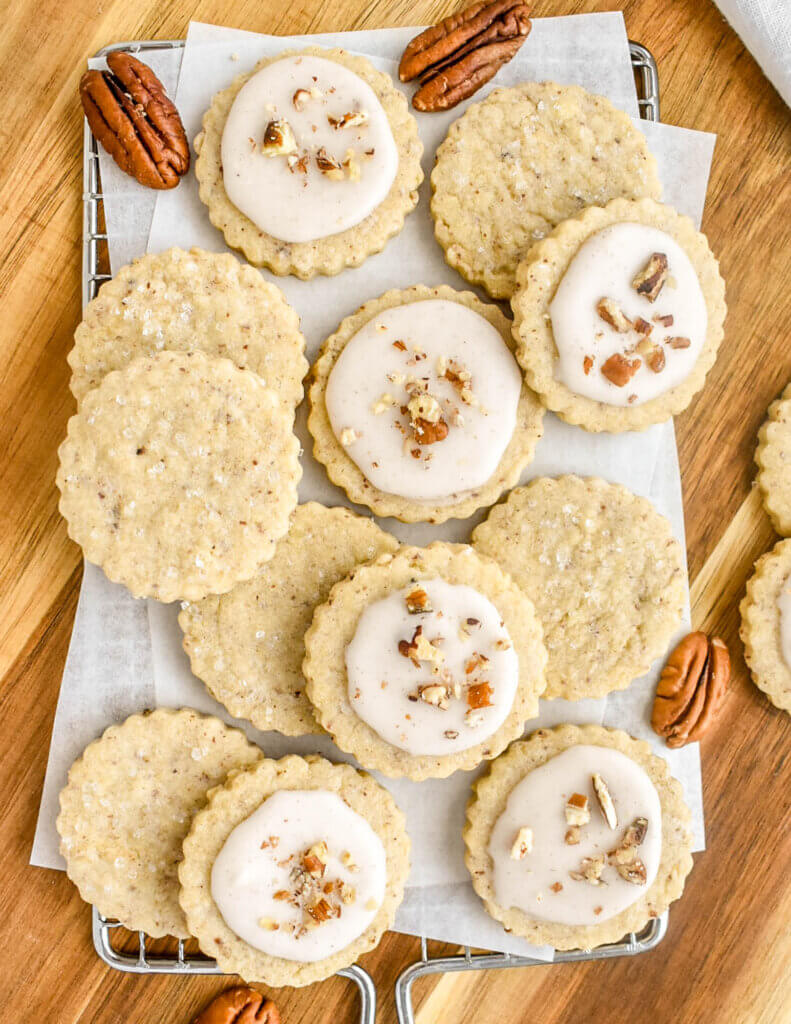 Pecan cookies, some with icing glaze and chopped pecans on them and some with coarse sugar sprinkled on them set on a parchment lined rack with pecan halves set around them.