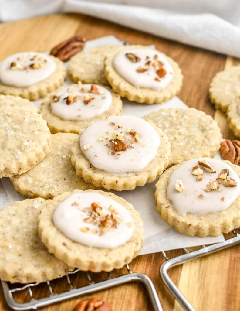 A pile of pecan cookies, some with icing and chopped pecans on them and some with coarse sugar sprinkled on them, set on a parchment lined rack.