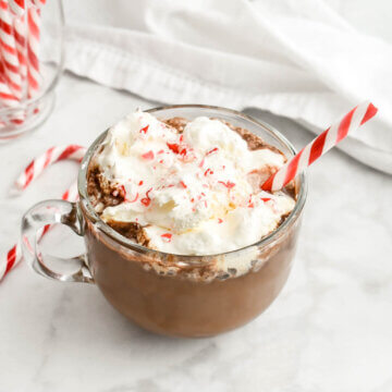 A peppermint mocha in a clear mug topped with whipped cream, crushed candy cane and a candy cane stick set on a marble countertop with a white napkin and candycanes around it.